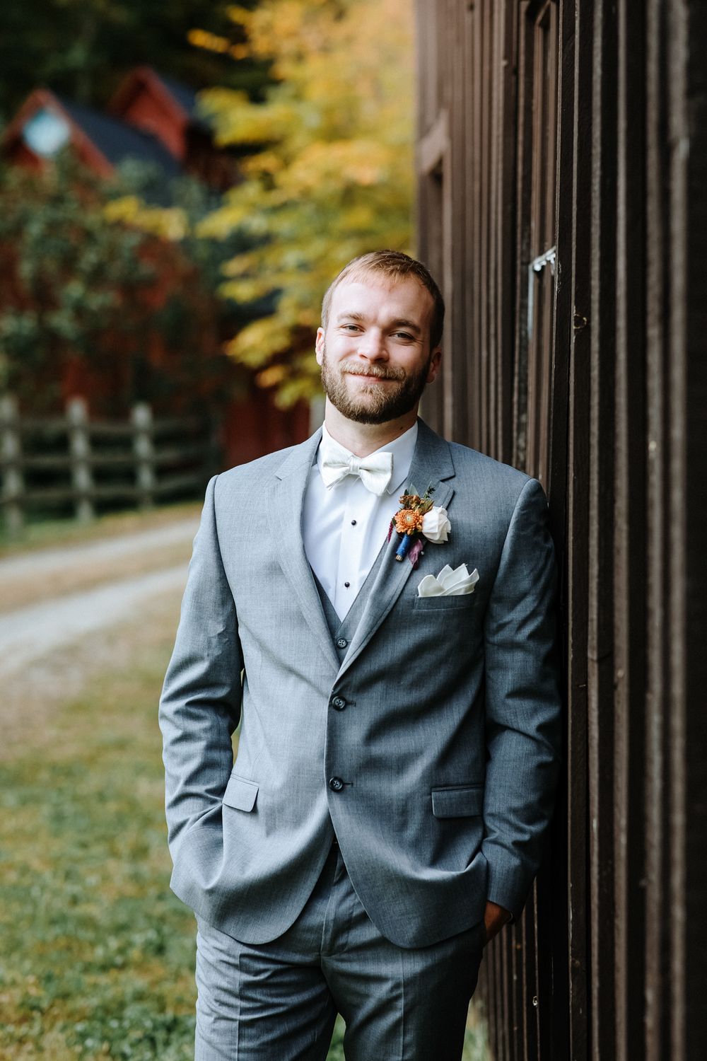 Blond groom with gray suit leans on wooden farm building with foliage in background for portrait. Riverledge Farm Grafton, VT wedding.