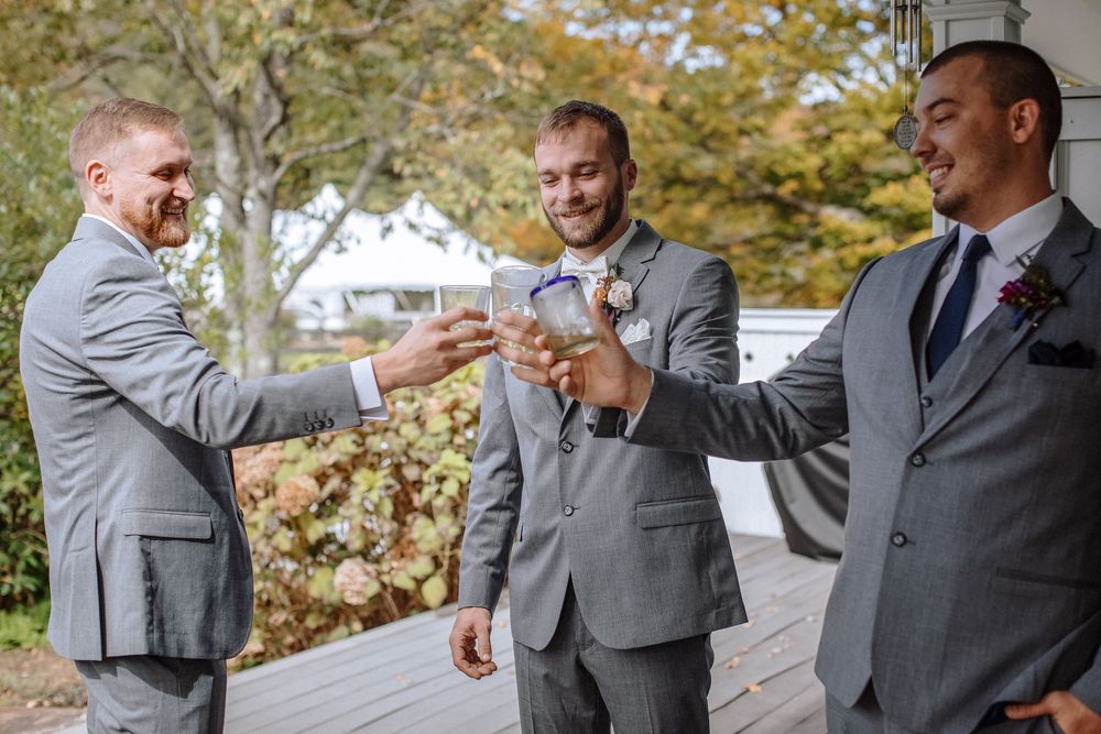 Three groomsmen in gray suits make a toast with an alcoholic drink on a gray porch on wedding day.  North Shore wedding photographer.