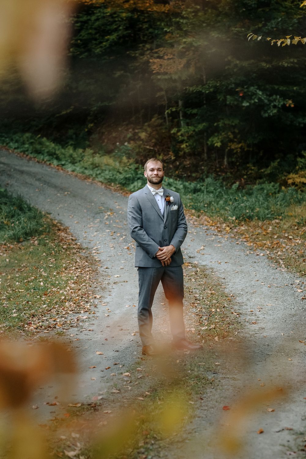Groom in gray suit with boutonniere stands on dirt path surrounded by autumn foliage at Riverledge Farm in Grafton, Vermont.