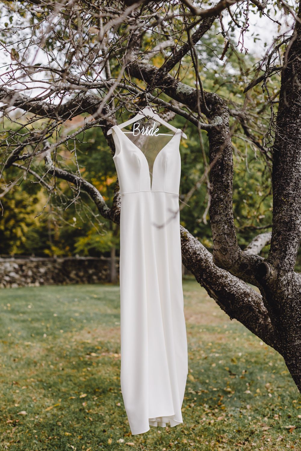 Elegant white slim fitting bridal gown hangs on a Bride hanger from a tree branch. North Shore MA Seacoast NH wedding photographer