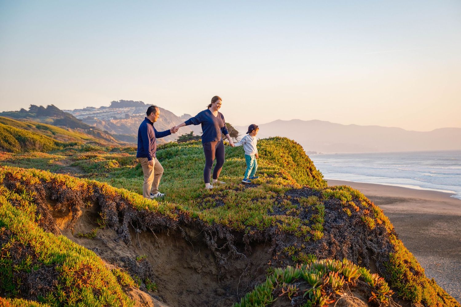 Family of three holding hands while hiking on lush green coastal hills at golden hour.