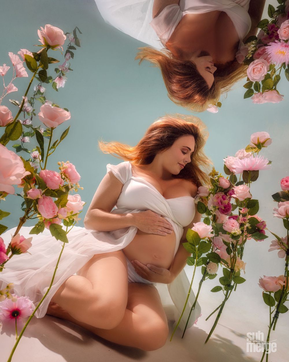 Artistic maternity photoshoot featuring pink hollyhock flowers and soft ethereal lighting in a dreamy studio setting.