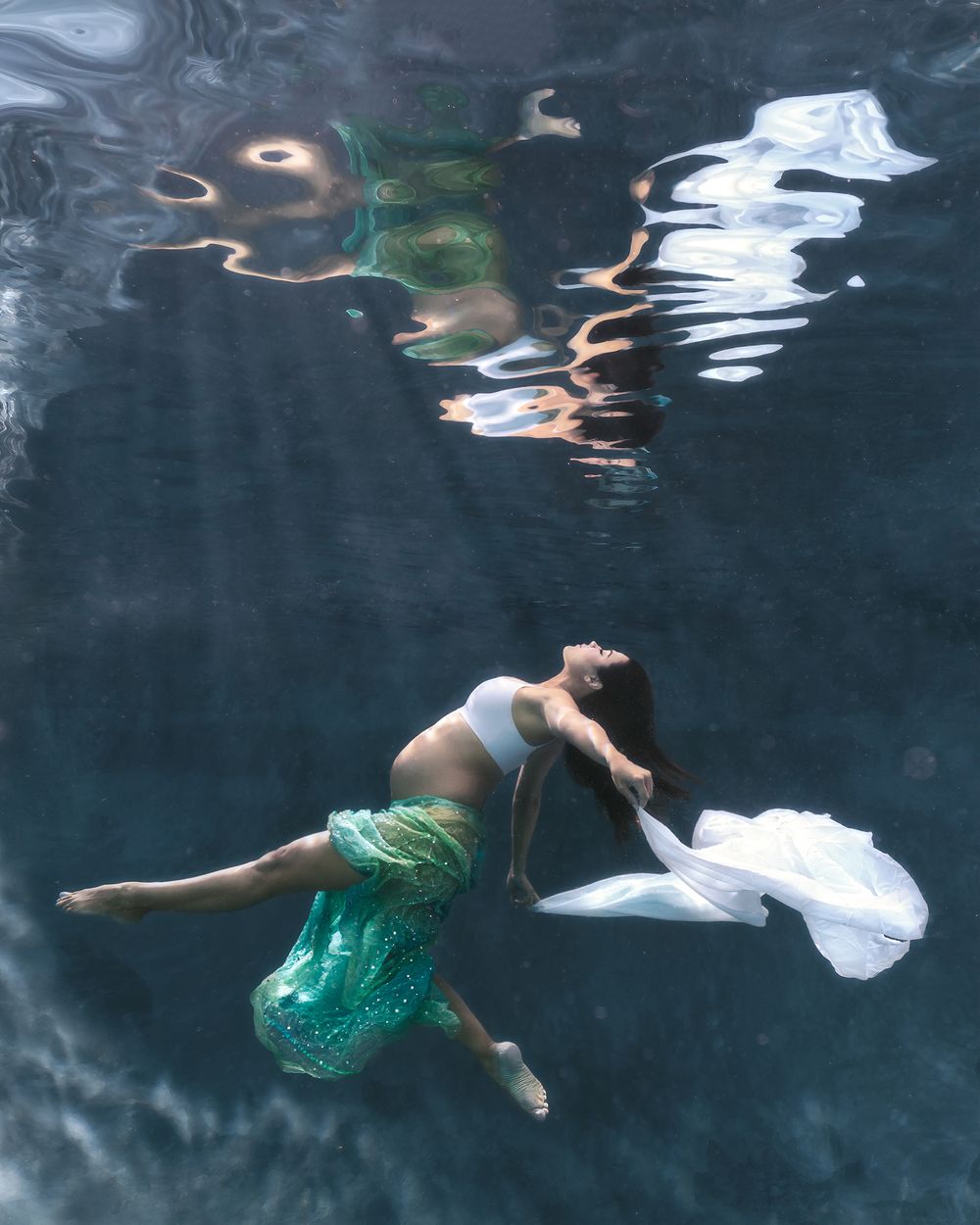 Underwater view of a figure in a green dress floating gracefully in dark blue water creating ethereal reflections.