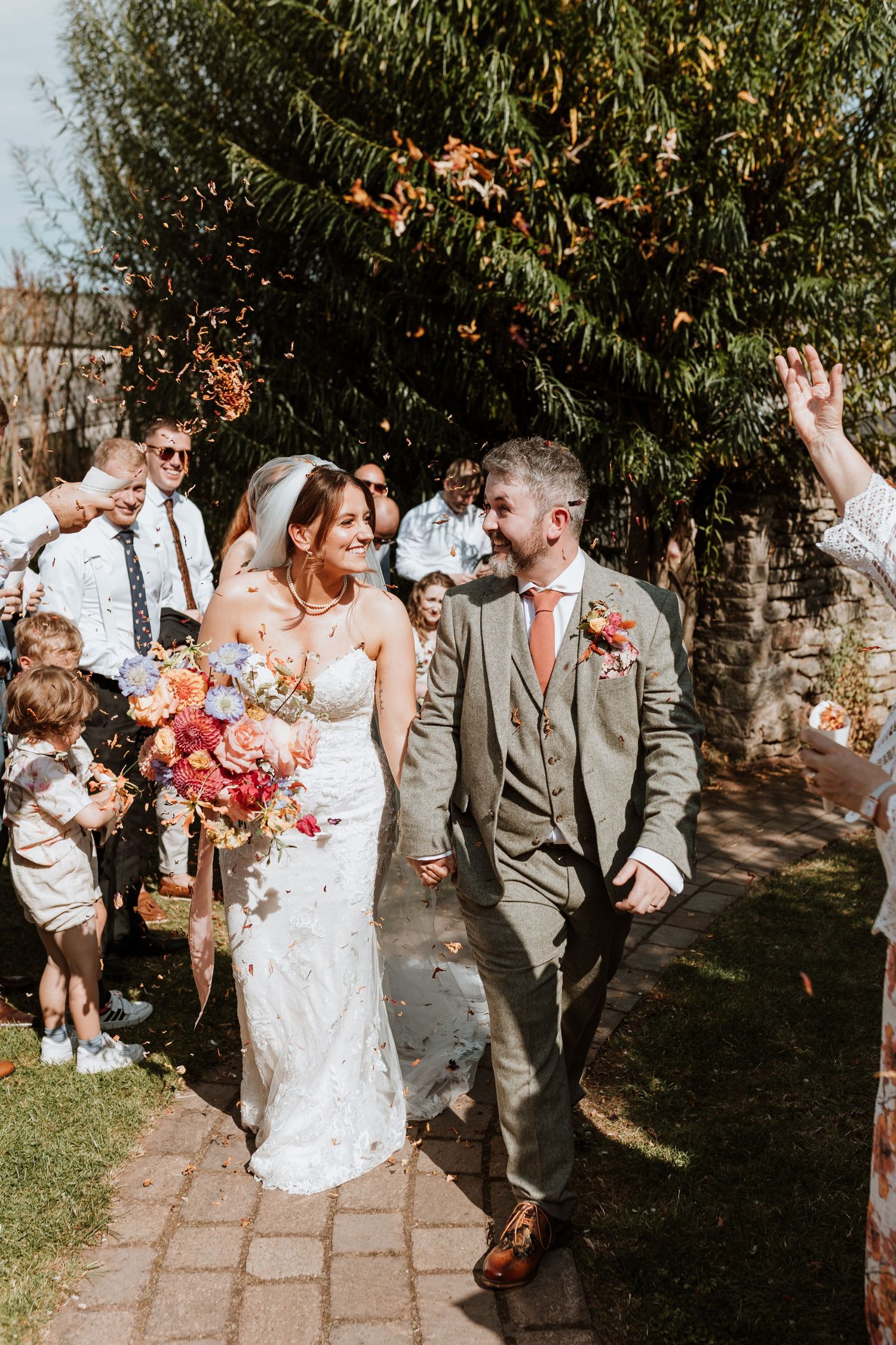 A joyful wedding celebration in a garden setting with guests throwing confetti as the newlyweds walk down a stone path.