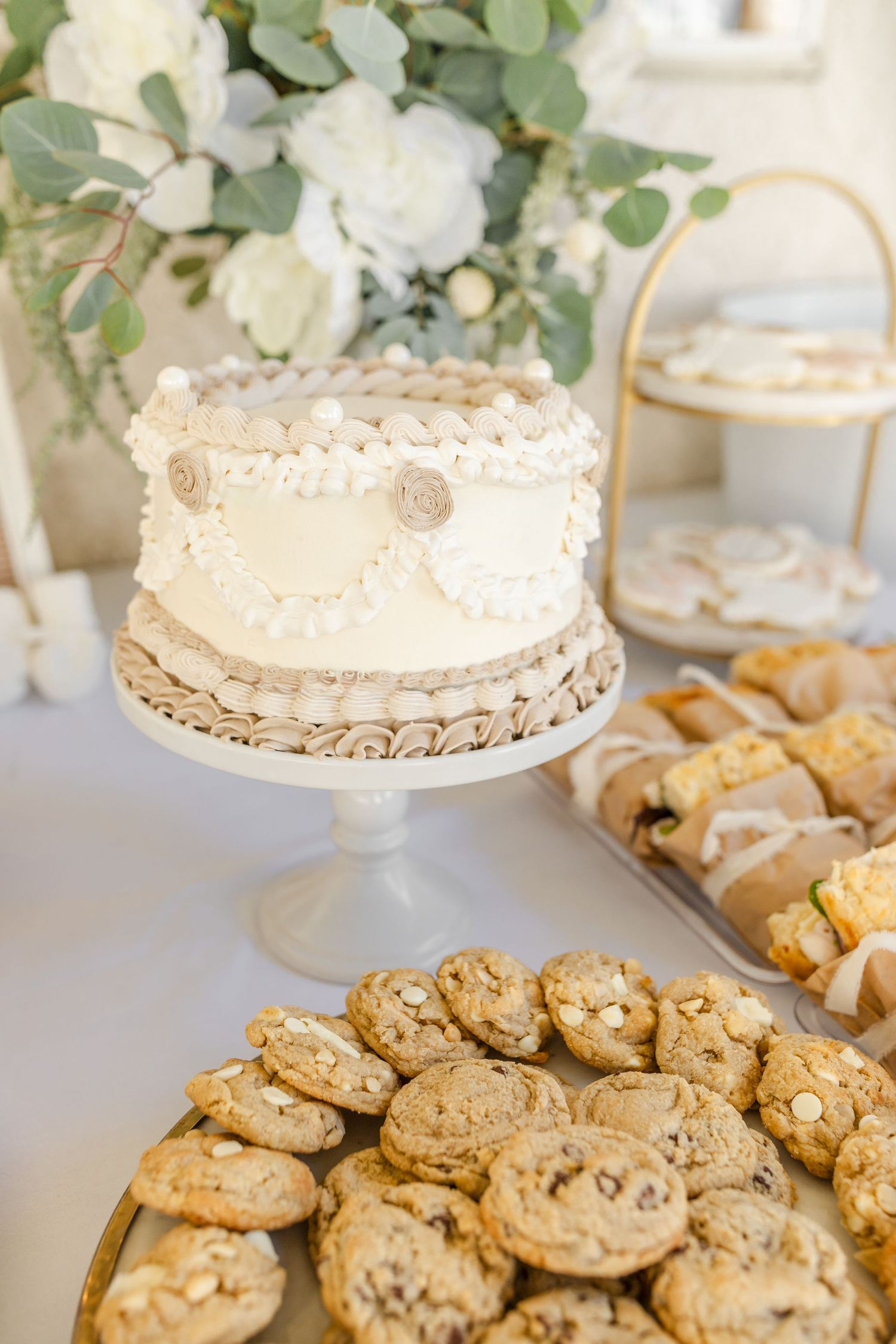 A white frosted wedding cake sits on a pedestal stand alongside golden brown chocolate chip cookies on a table.