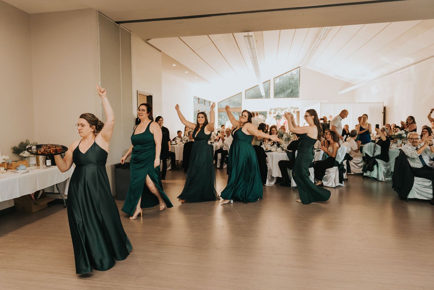 Wedding guests in dark teal bridesmaid dresses dance together on a white dance floor at an elegant reception.