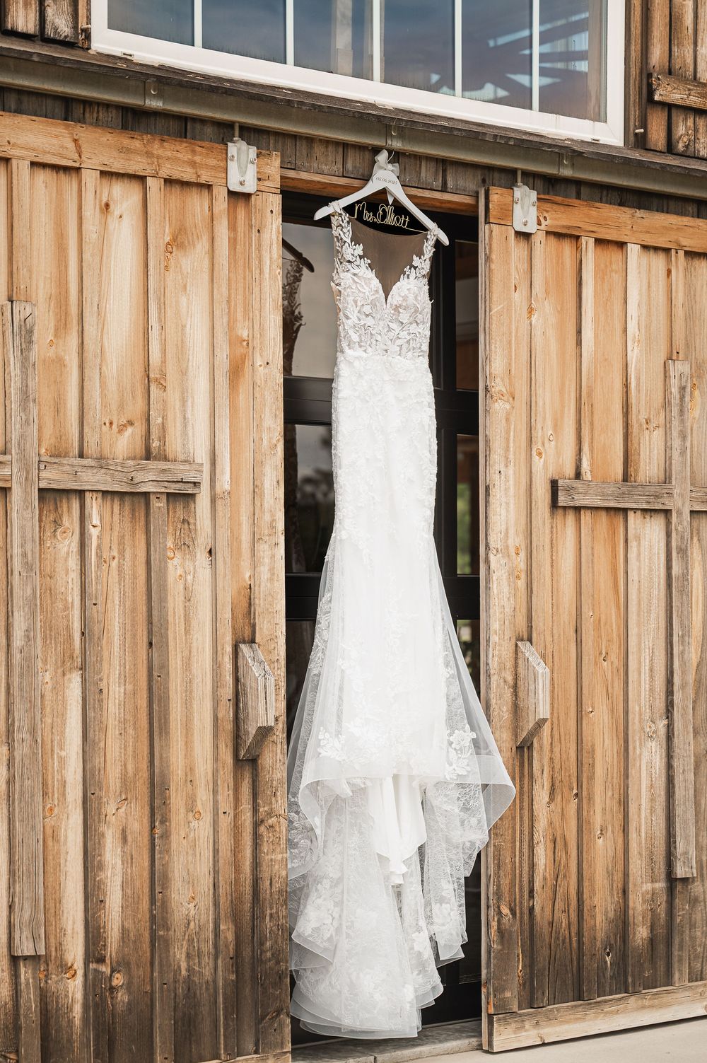 Elegant white lace wedding dress hanging on wooden barn door exterior.