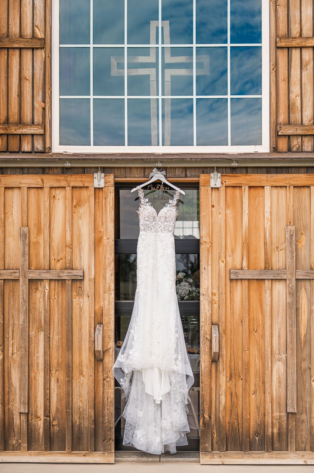 Wedding gown displayed against rustic wooden barn with cross visible through window.