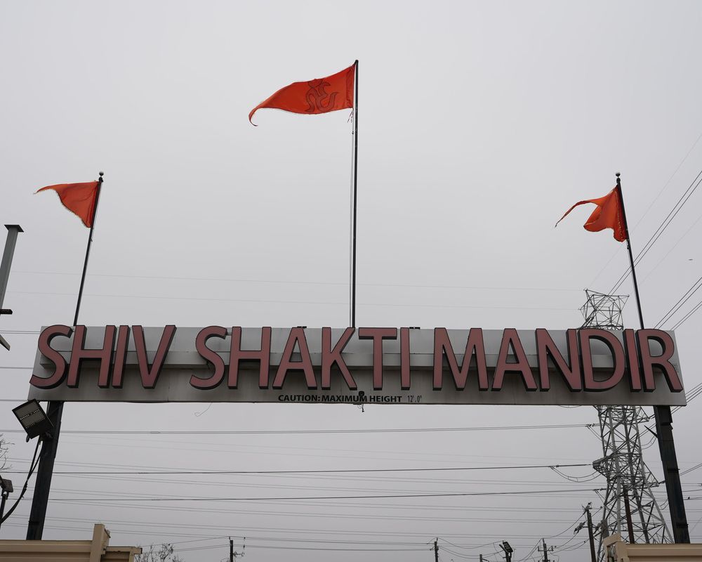 Red flags wave in front of a sign reading 'SHIV SHAKTI MANDIR' against an overcast sky.