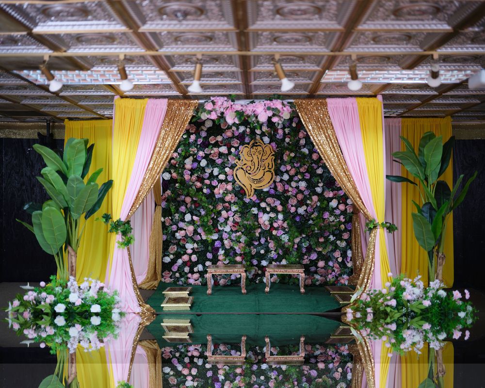 Elegant wedding mandap decorated with pink and white flowers, yellow drapes, and green foliage against a floral backdrop.