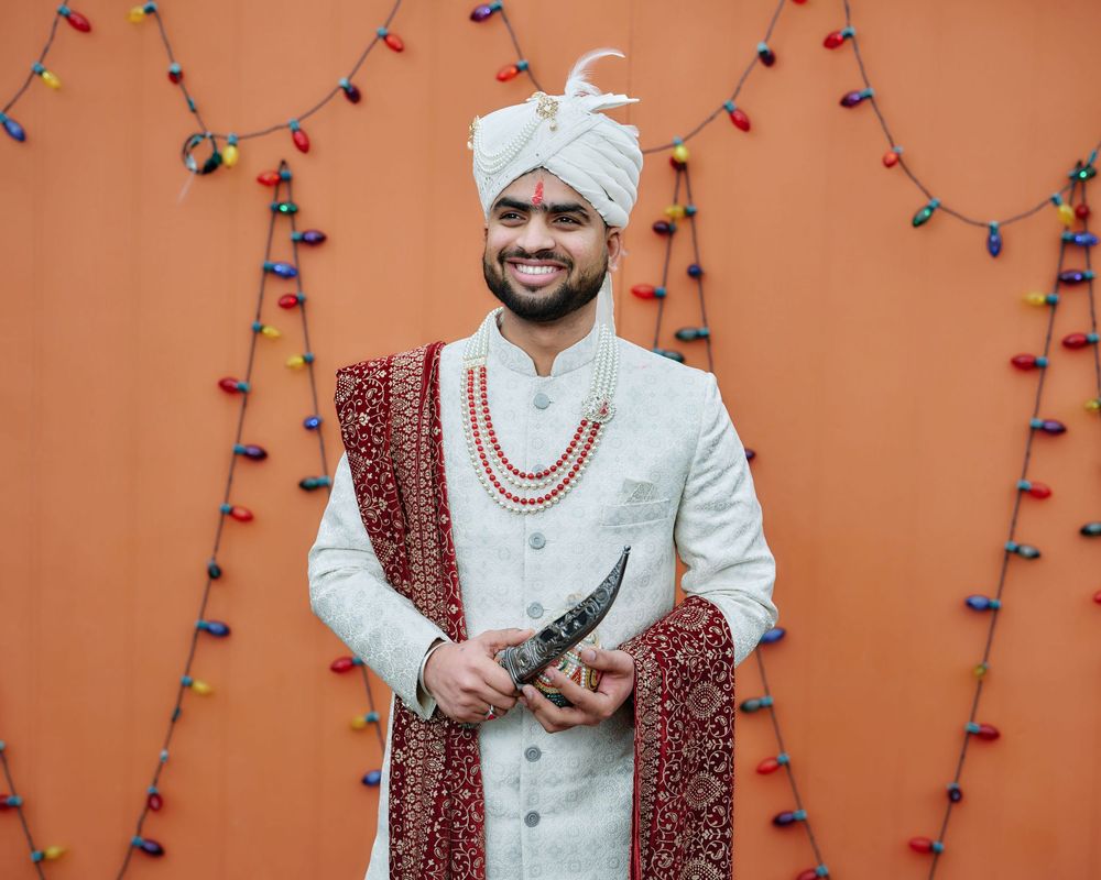 Traditional Indian groom in white sherwani and maroon dupatta poses against orange wall with festive string lights.