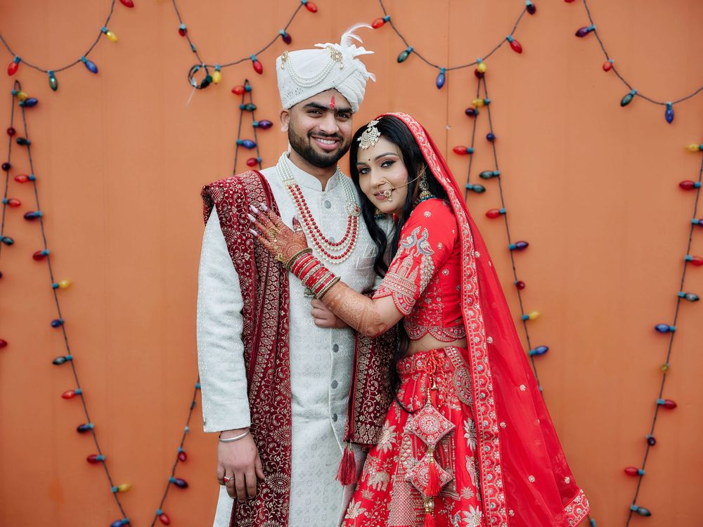 A couple in traditional Indian wedding attire poses against a terracotta wall with string lights.