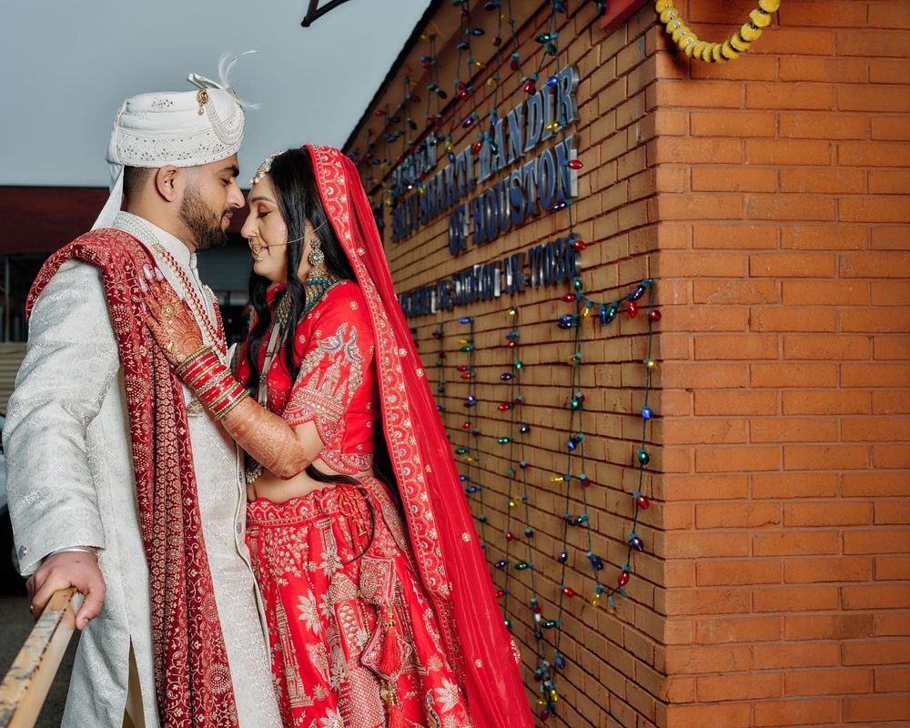 Traditional Indian wedding couple in red and white attire posing against a brick wall backdrop.