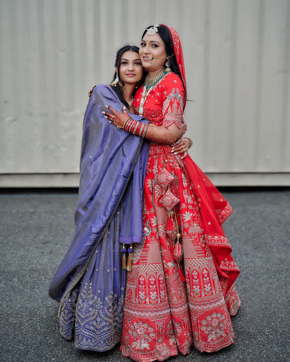 Two people in traditional Indian wedding attire, one in purple and one in red, embrace during a photo session.