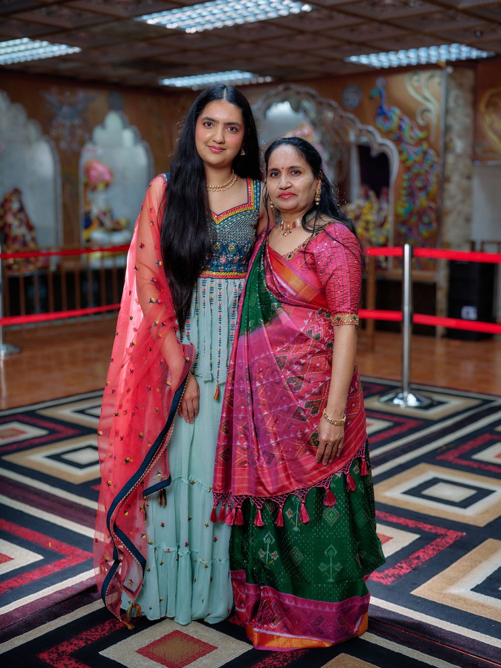 Two people in traditional Indian attire pose together on a patterned floor with geometric designs.