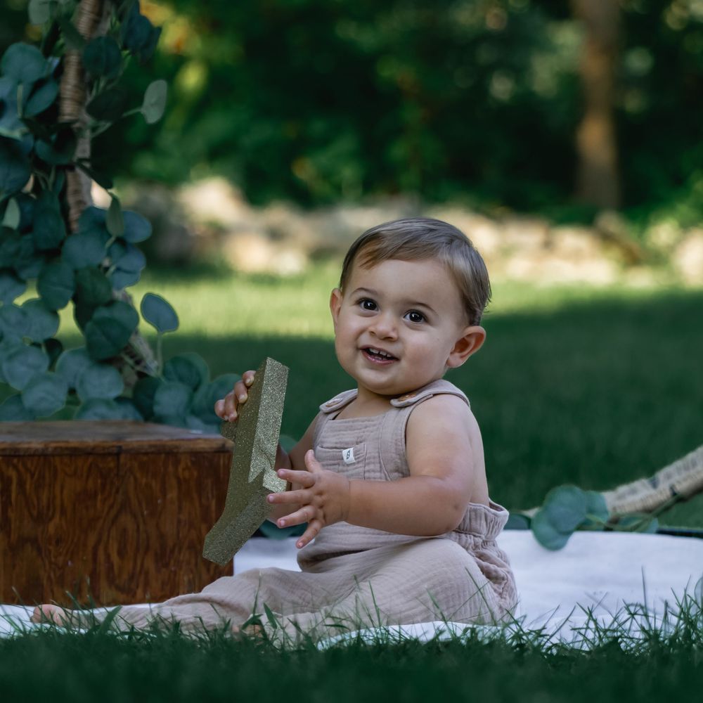 Smiling baby in linen outfit sitting on blanket in garden with eucalyptus decor.