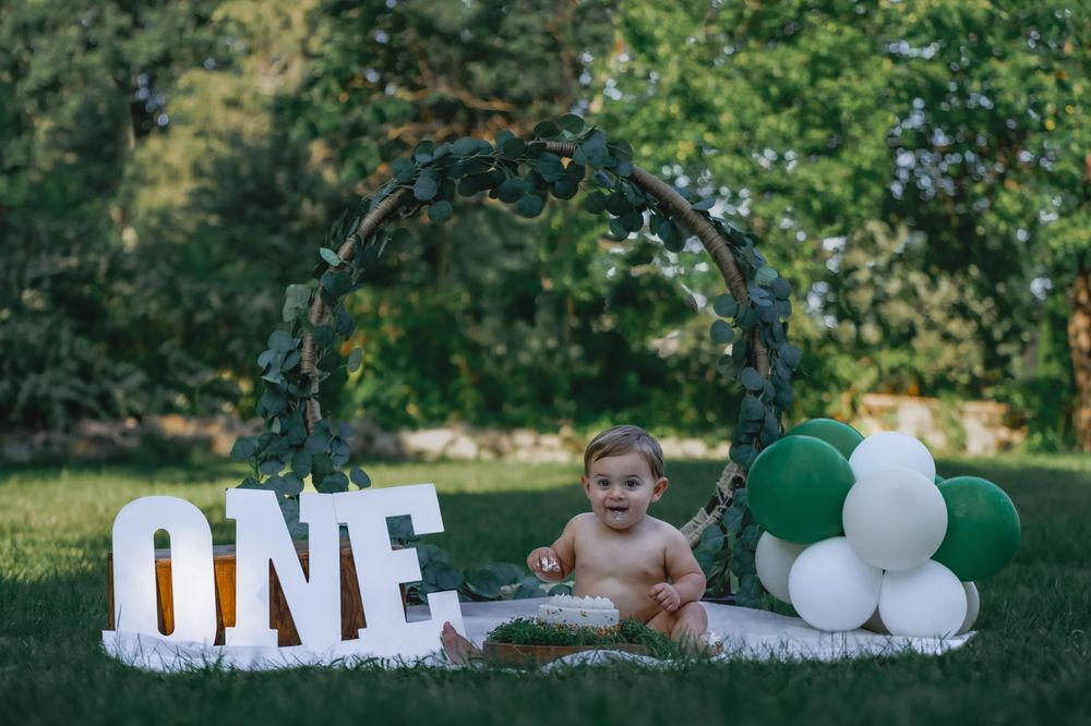 Baby sitting near ONE sign with green and white balloons during first birthday celebration.