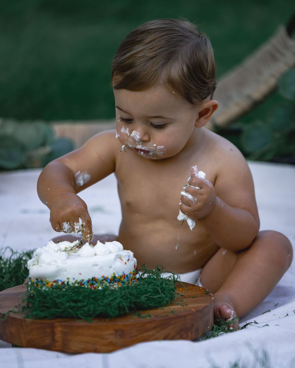 Baby exploring first birthday cake smash outdoors with white frosting on face.