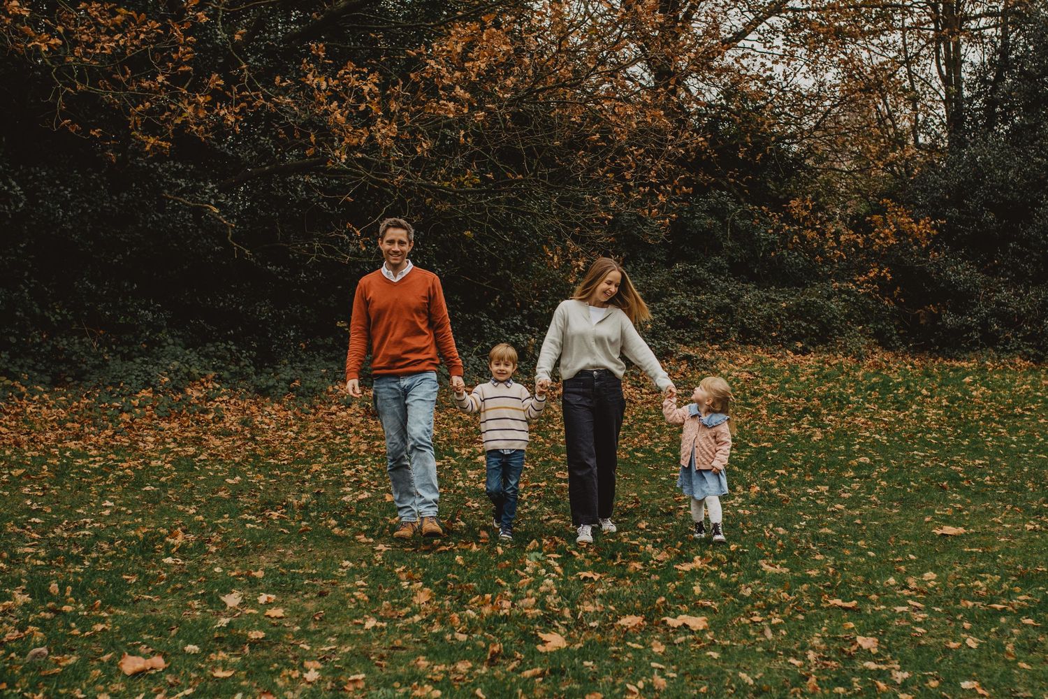 Family walks together through autumn park with fallen orange leaves scattered across grass on cloudy day.