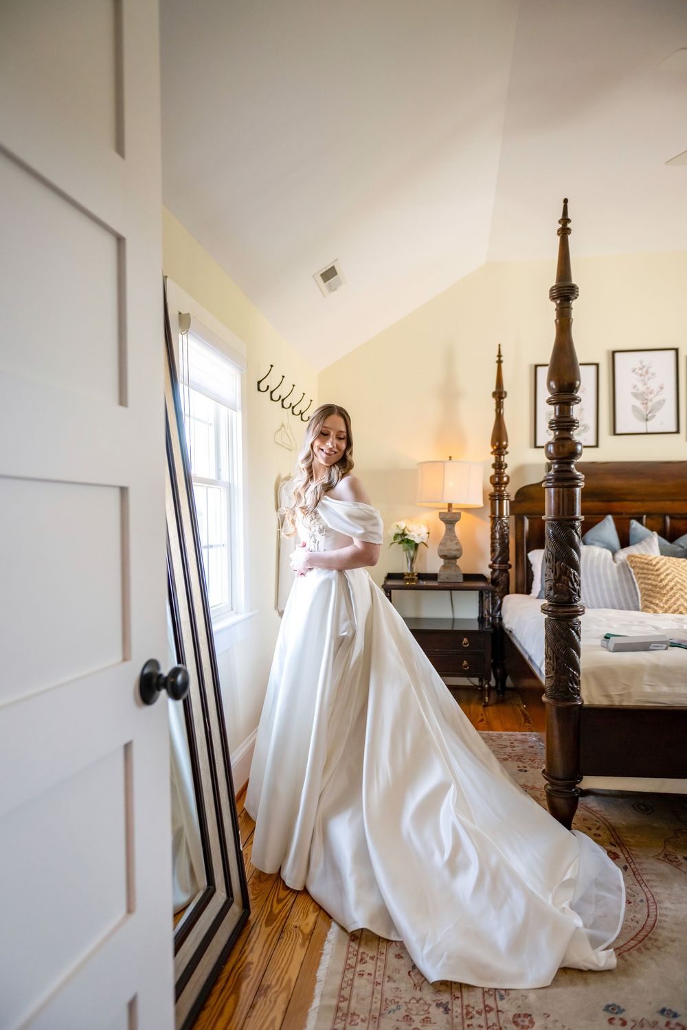 A bride in an elegant white satin ball gown stands in a sunlit bedroom with a four-poster bed and hardwood floors.