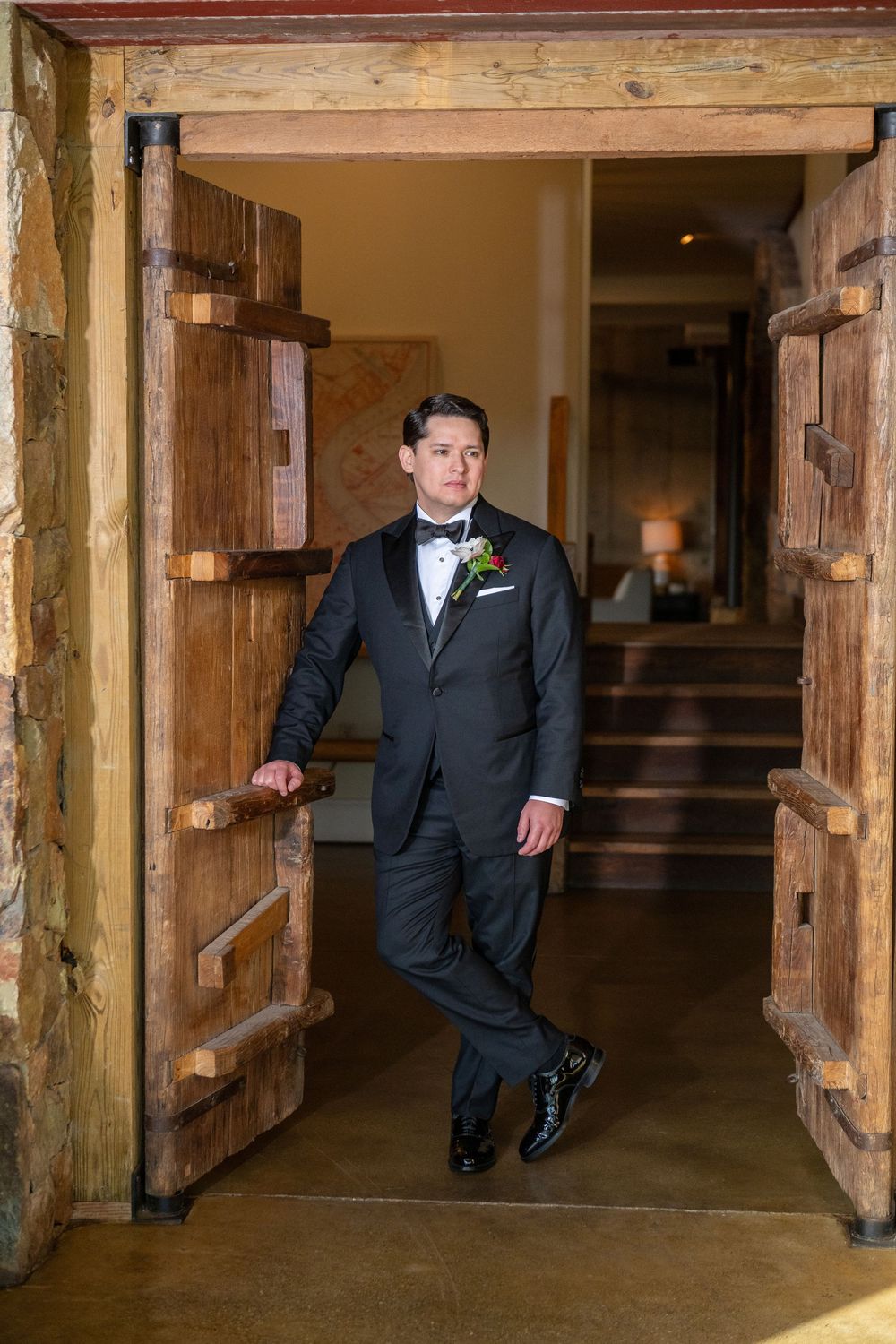 A groom in a classic black tuxedo leans against a rustic wooden doorframe in an elegant venue hallway.