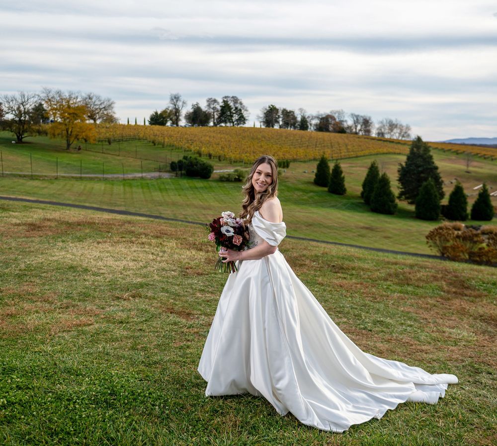 A bride in a flowing white gown stands in a scenic autumn field holding a bouquet.