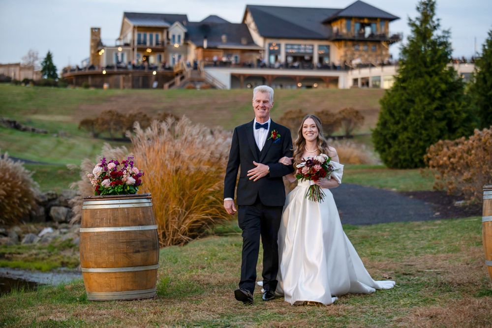A wedding couple poses in front of a grand estate with a rustic wine barrel and autumn foliage.