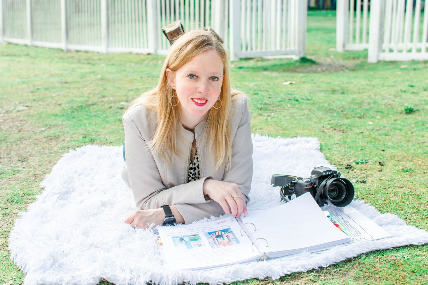 A photographer in a bright red cardigan and glasses crouches near vibrant red roses in an outdoor garden setting.