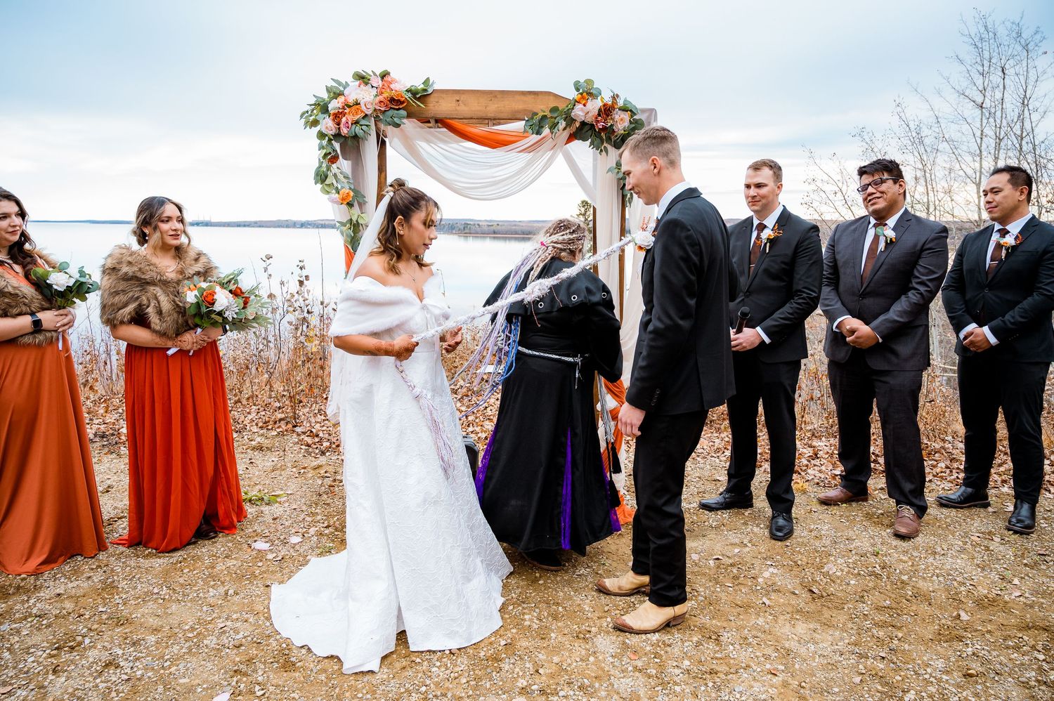 A fall wedding party poses outdoors with bridesmaids in burnt orange dresses and groomsmen in dark suits.