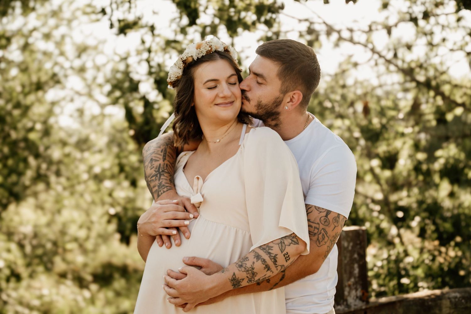 Couple with tattoos embracing outdoors, man kissing pregnant woman wearing white dress.