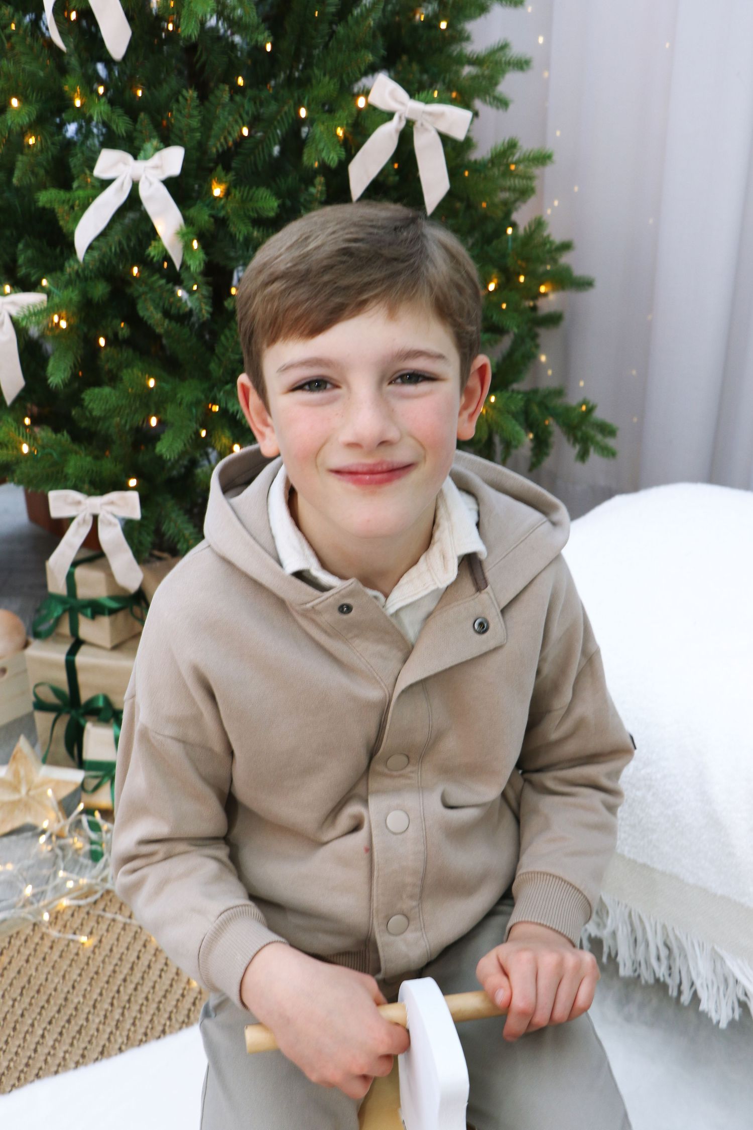 A young child poses in a beige jacket next to a decorated Christmas tree with white bows.