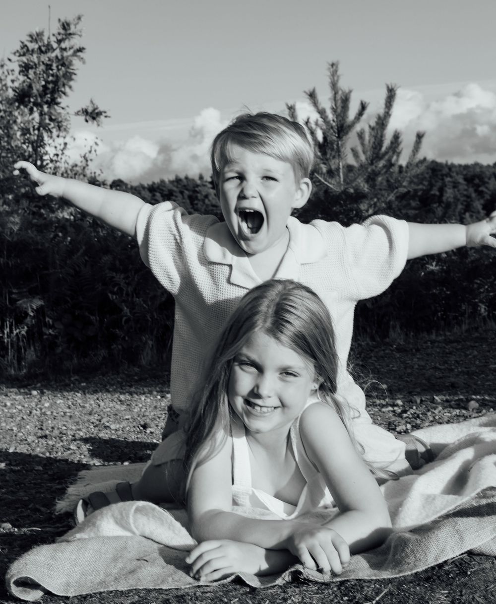 Black and white photo of a young girl lying down on a blanket smiling with her brother sitting on her back laughing with his arms out.