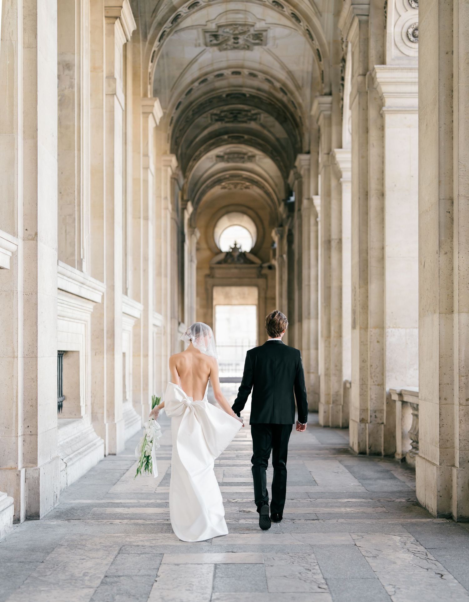 A couple walks through an elegant arched colonnade hallway with classical architecture and natural lighting.
