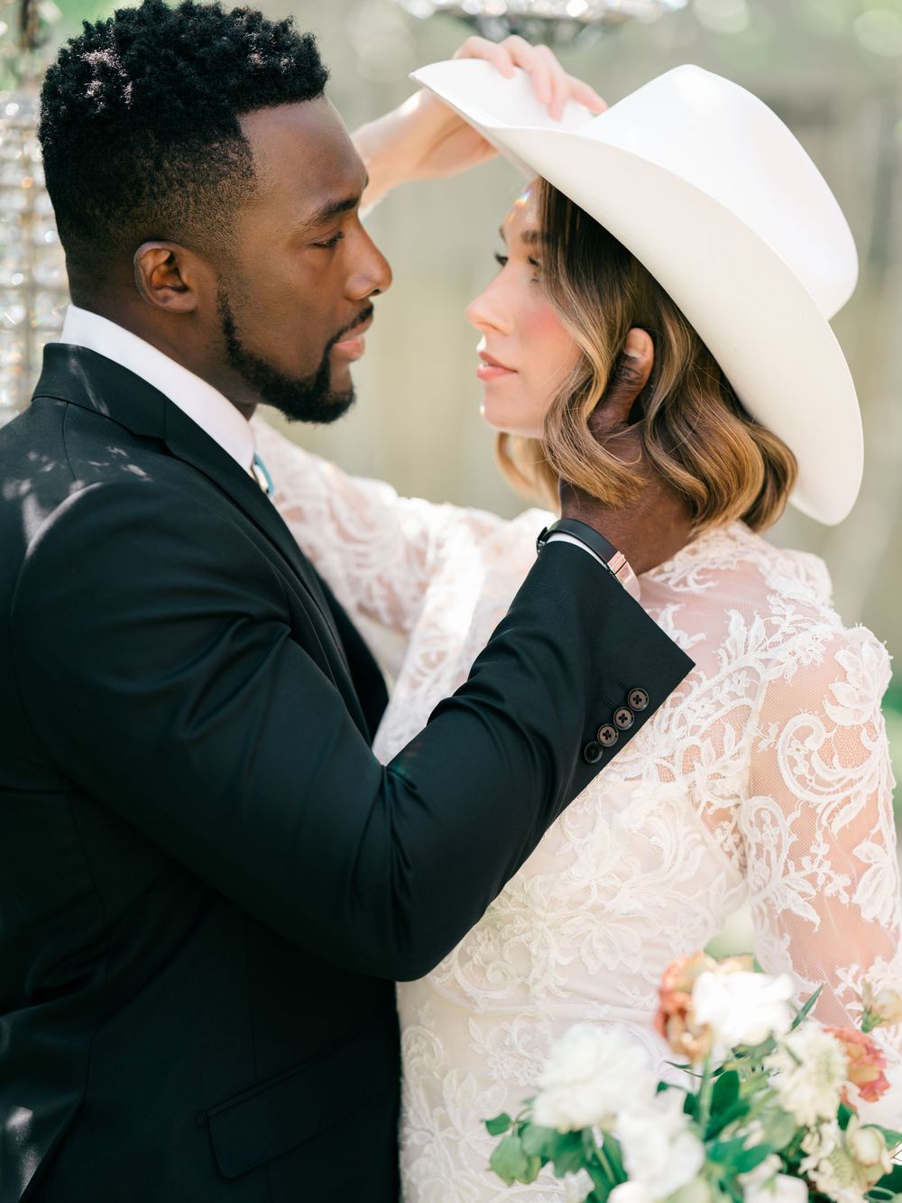 A romantic wedding portrait in soft focus showing a couple in formal attire with white hat and floral bouquet details.
