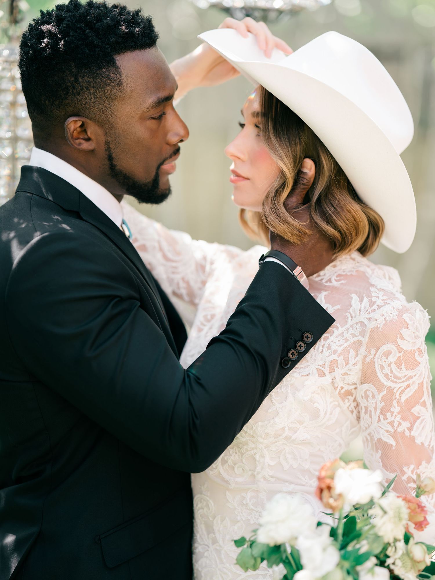 A romantic wedding portrait in soft focus showing a couple in formal attire with white hat and floral bouquet details.