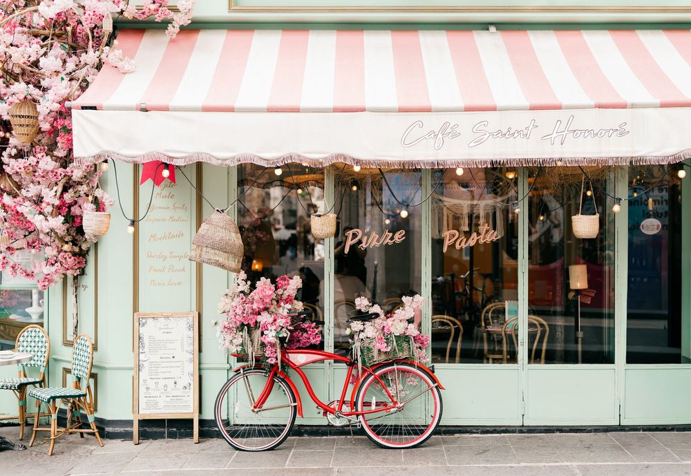 Pink cherry blossoms frame a row of charming storefronts with striped awnings and a vintage bicycle parked outside.