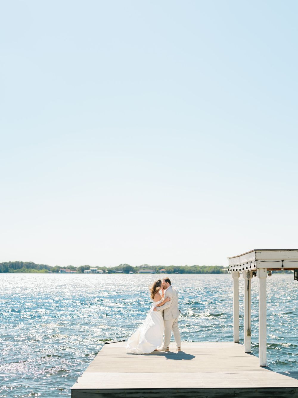 A couple shares a romantic moment on a wooden pier overlooking sparkling blue waters on a sunny summer day.