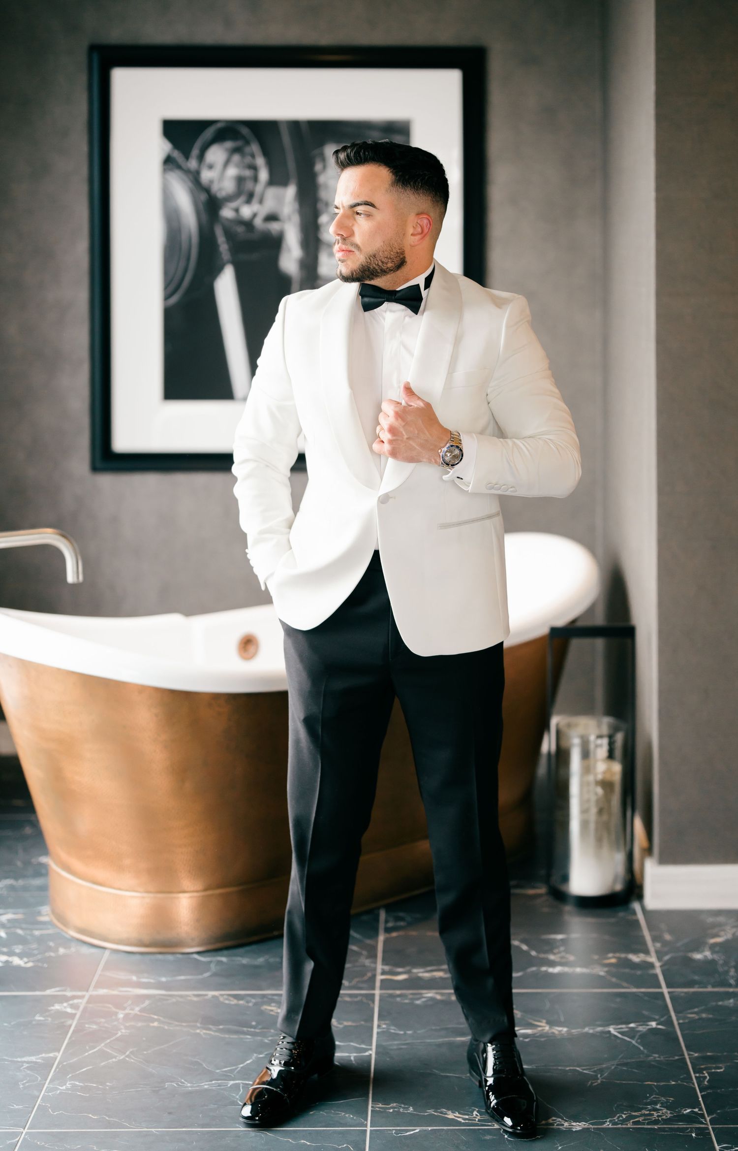 Elegant groom in white tuxedo jacket poses in modern bathroom with copper tub.