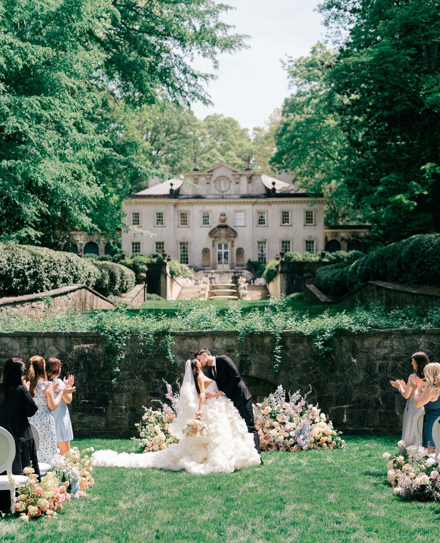 Bride and groom share a kiss at an elegant outdoor garden wedding ceremony with floral arrangements and a grand estate backdrop.