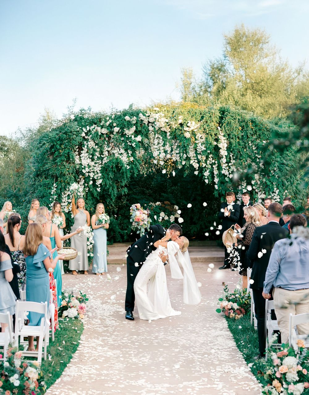 A panoramic view of an elegant wedding ceremony with guests gathered under floral arches in an outdoor garden setting.