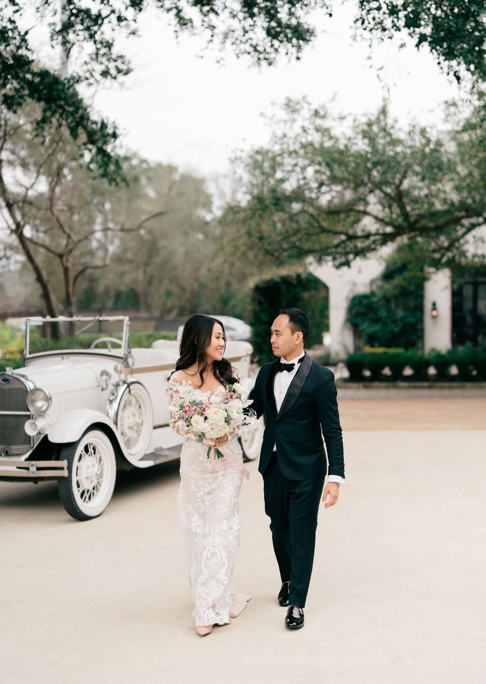 A couple in formal attire share a romantic moment by a vintage car in a dreamy outdoor setting.