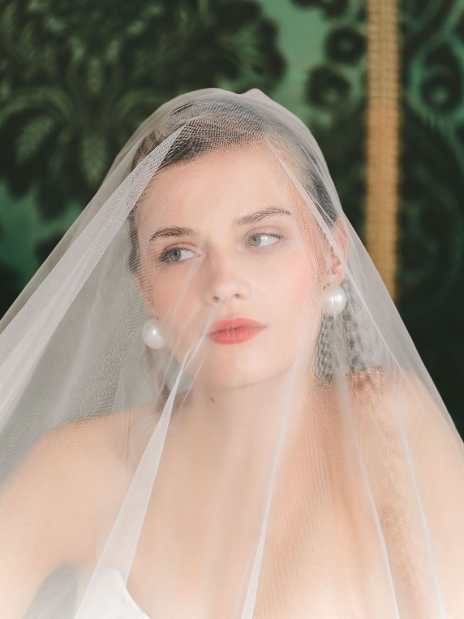 An elegant bridal portrait featuring a delicate tulle veil and pearl earrings against a garden backdrop.