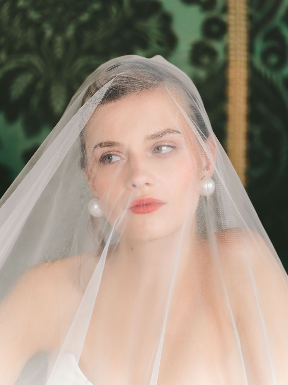 An elegant bridal portrait featuring a delicate tulle veil and pearl earrings against a garden backdrop.