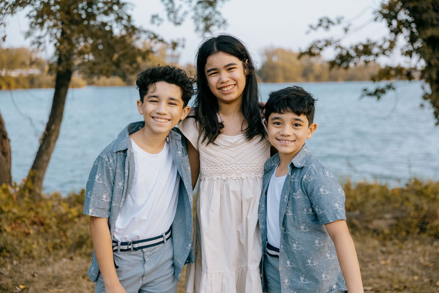 Three siblings smile together while standing by a lake on an autumn day with trees in the background.