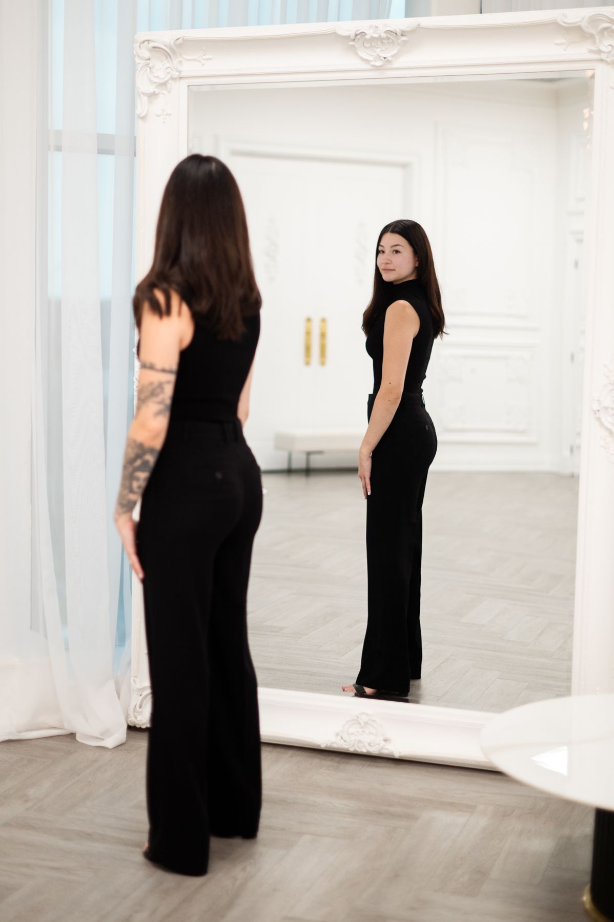 Multiple reflections in mirrors show figures in black dresses within a white gallery space.