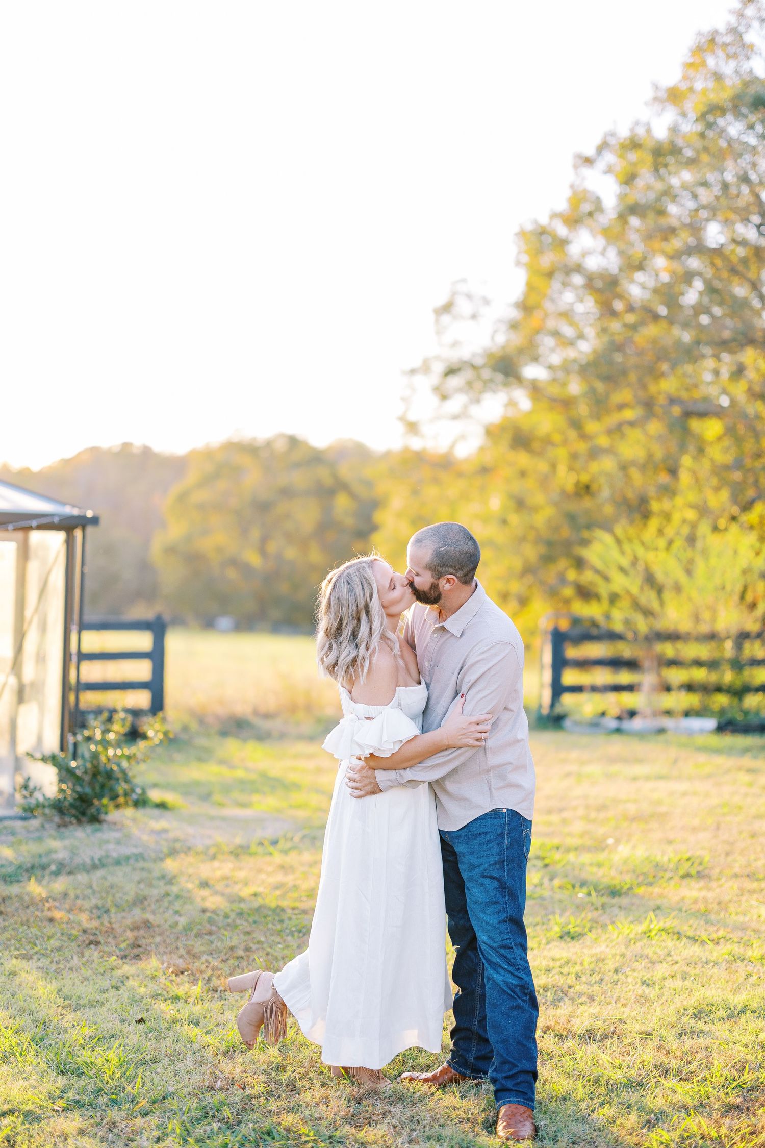 Romantic couple embraces in golden sunlight on rustic farm property with wooden fence and trees in background.