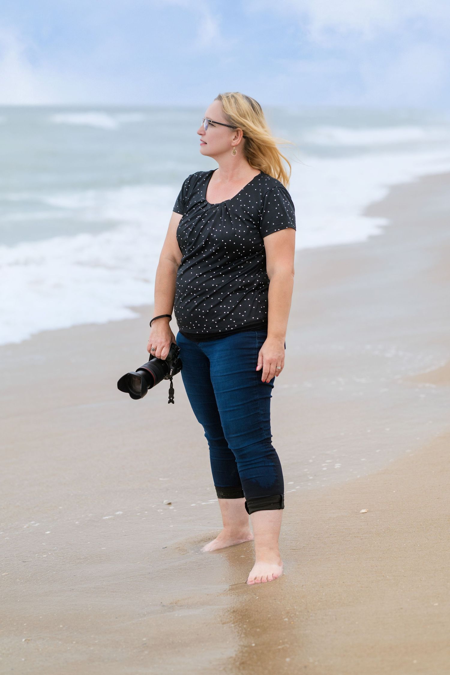 Photographer walking along beach shoreline with camera in casual attire.