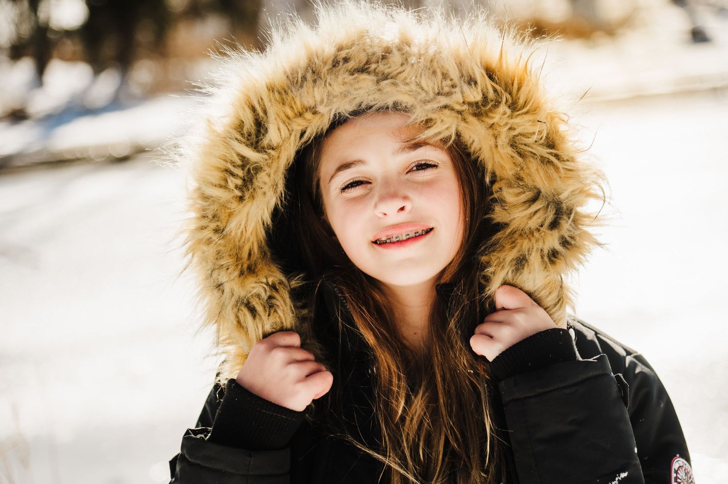 A winter portrait of someone wearing a fur-trimmed hood coat in snowy outdoor setting with soft bokeh lighting effects.