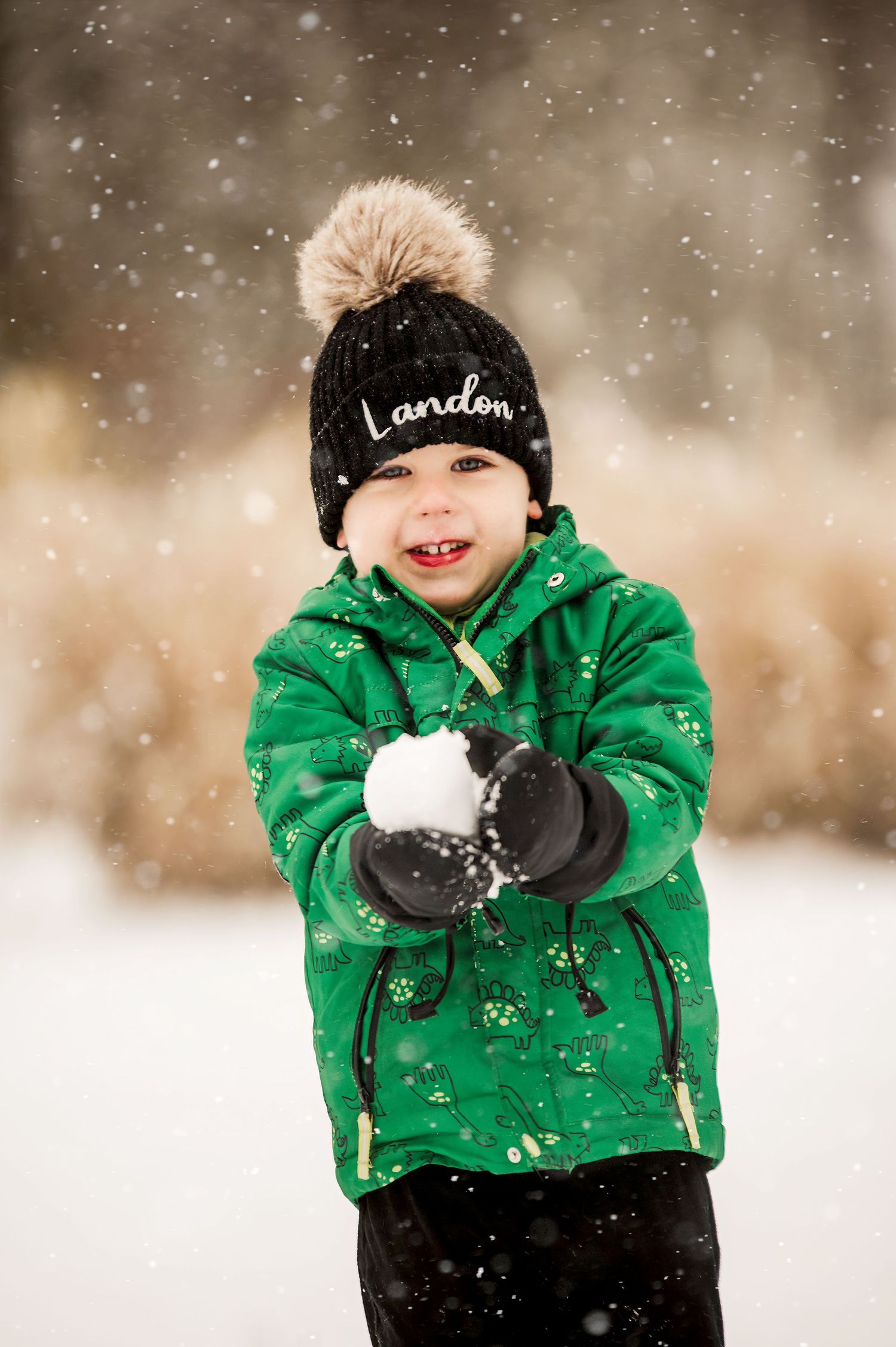 A child wearing a green winter coat and black beanie plays in the falling snow while holding a snowball.