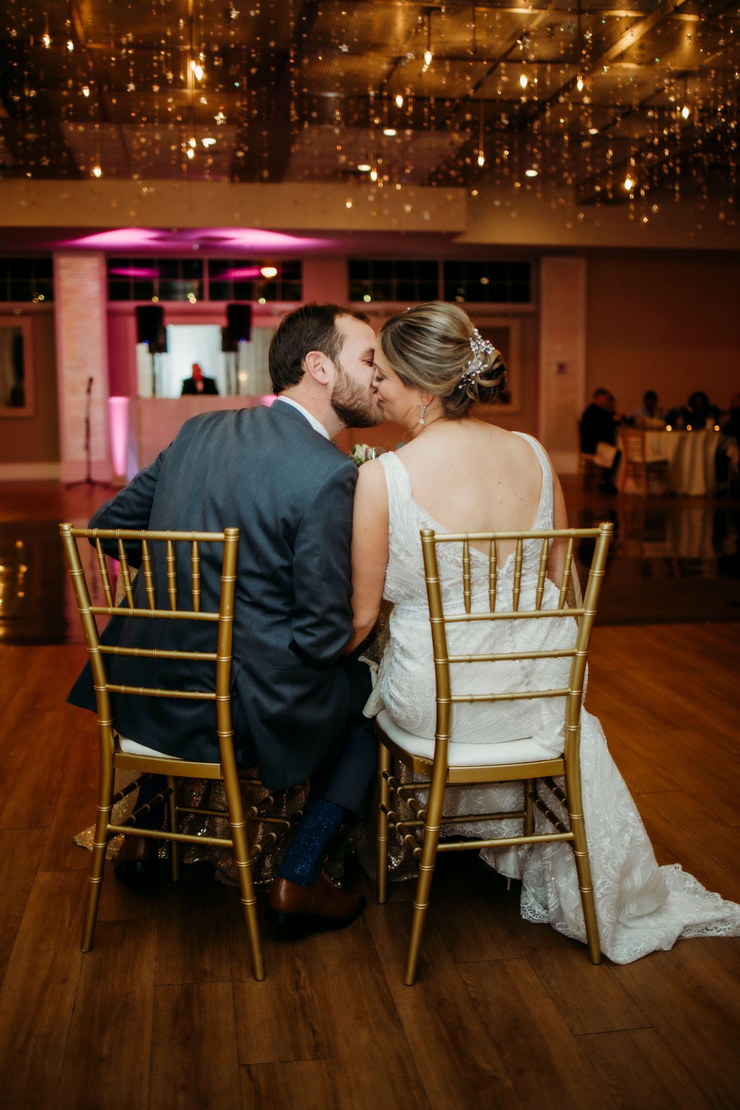 A romantic couple sits together on gold chairs under twinkling lights at their wedding reception.